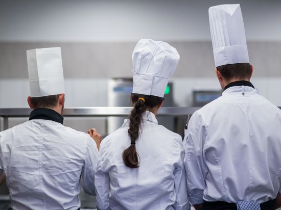 Drei Jugendliche stehen mit Kochmütze und Kochjacke in einer Restaurantküche.