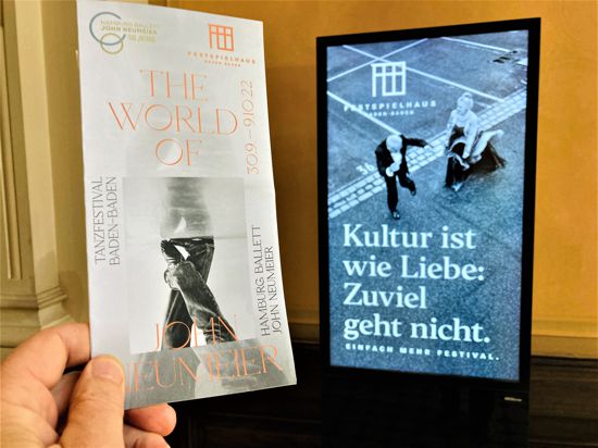 Blick auf eine Videotafel neben dem Ticketverkauf des Festspielhauses Baden-Baden im Alten Bahnhof. Daneben hält eine Hand einen Flyer zum bevorstehenden Tanzfestival. 