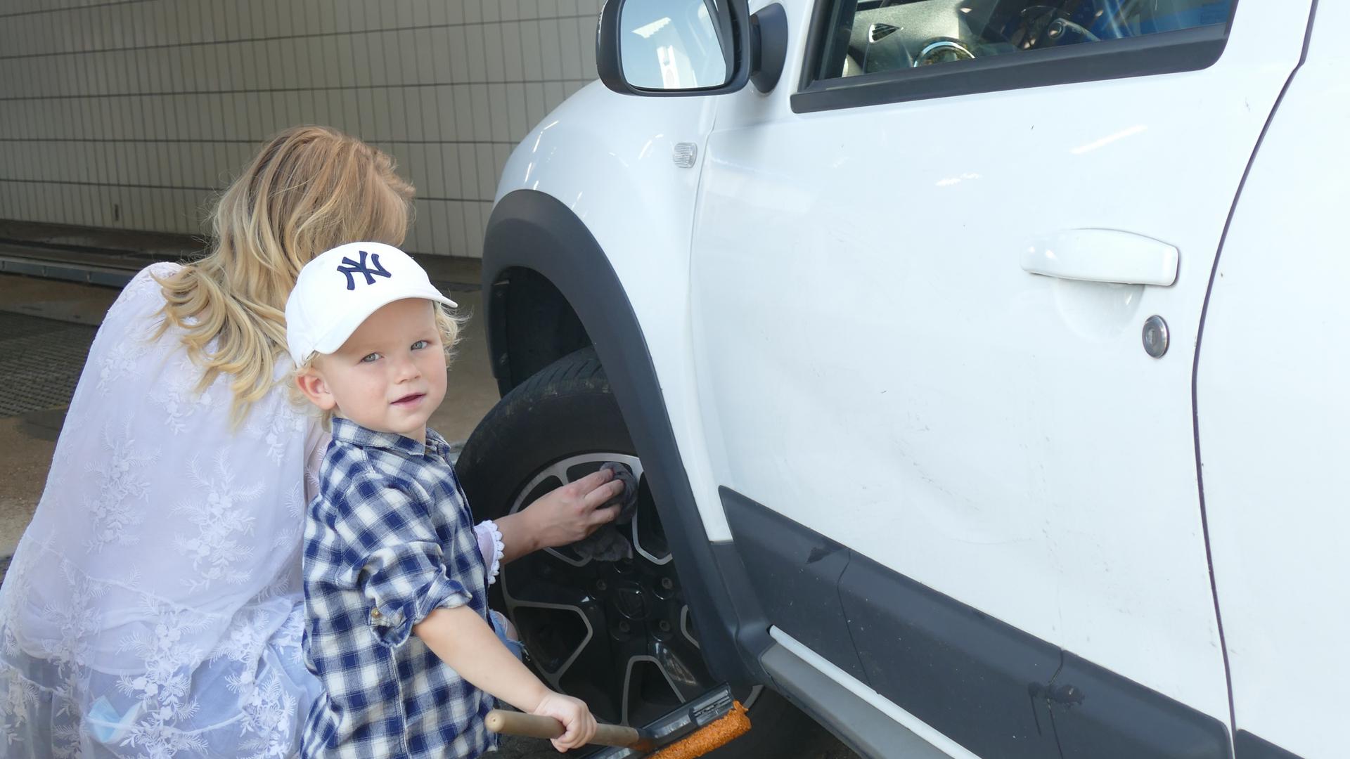 Eine Frau reinigt die Fegen von einem weißen Auto und ein Kleiner Junge steht mit einem Schbenreiniger in der Hand daneben.