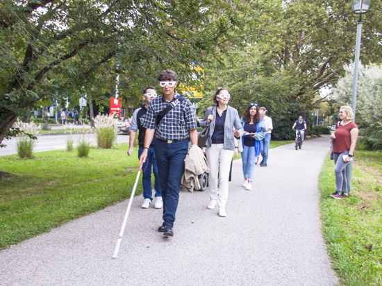 Eine Gruppe von Menschen läuft in Baden-Baden über einen Fußgängerweg. Sie haben einen Blindenstock in der Hand und tragen spezielle Brillen.