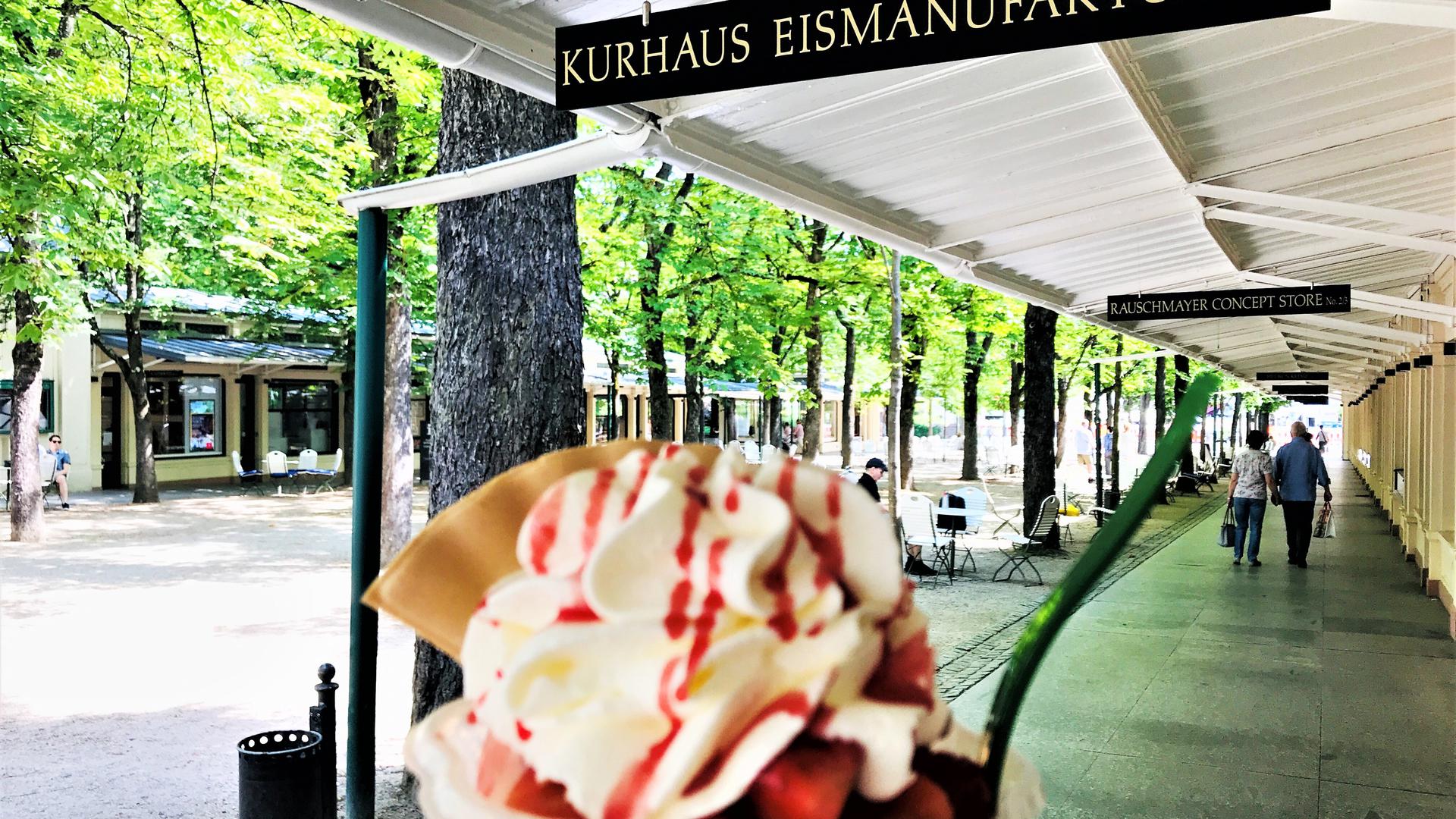 Ein Genießer hält einen Eisbecher ins Bild. Im Hintergrund sind die Kurhaus-Kolonnaden in Baden-Baden mit der Kastanienallee zu sehen. 