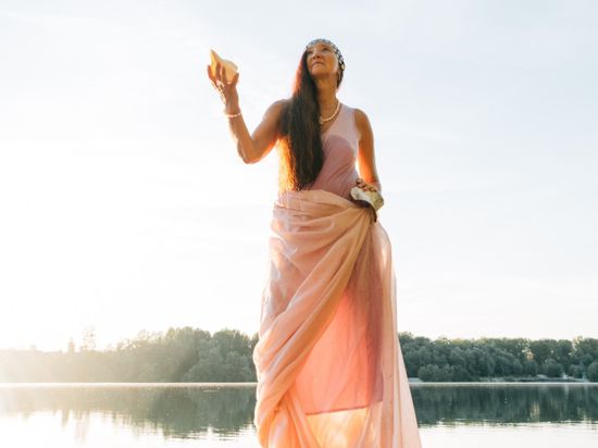 Sängerin Eva Weis steht im Kleid an einem See und hält eine Muschel in der Hand.