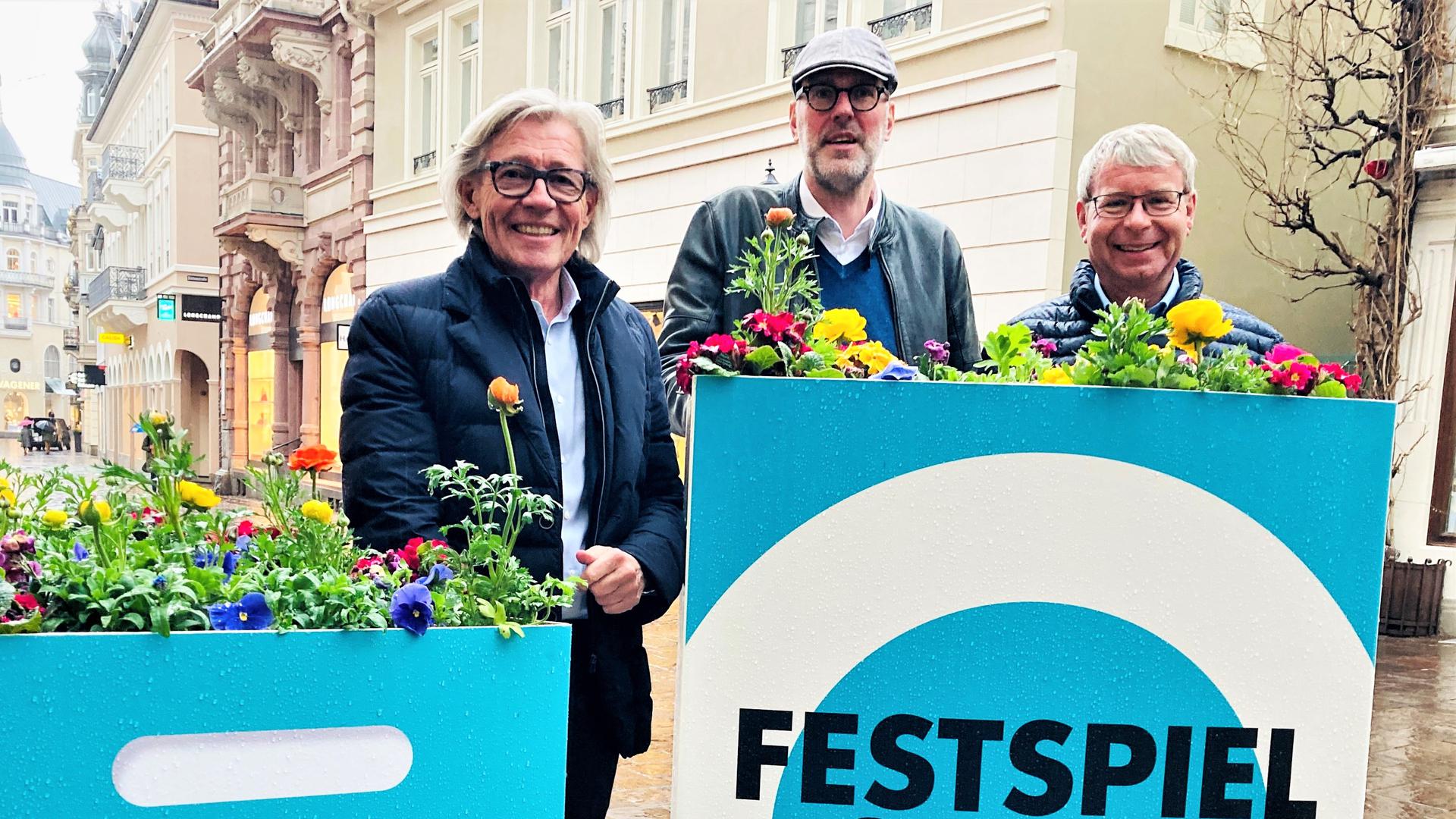 Einzelhändler Franz-Bernhard Wagener (von links), Festspielhausintendant Benedikt Stampa und Gartenbauamtschef Markus Brunsing stehen vor einer Blumentasche in der Fußgängerzone. 
