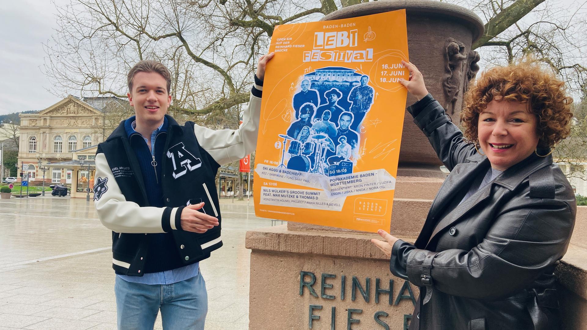 Nora Waggershauser und Konstantin Kölmel zeigen auf der Fieser-Brücke ein Plakat mit dem Programm des „Baden-Baden lebt“-Festivals.