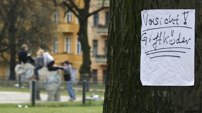 Ein Zettel mit einer Warnung „Vorsicht! Giftköder“ hängt an einem Baum in einem Park.