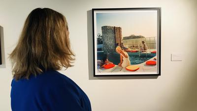 Eine Frau betrachtet ein Bild, auf dem Menschen am Rande eines Swimmingpools sitzen. Es ist ein Werk des amerikanischen Fotografen Slim Aarons.