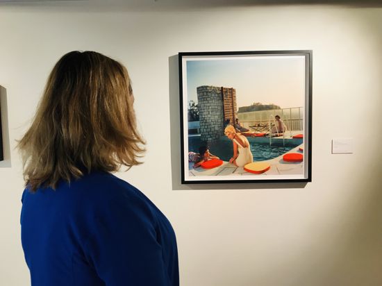 Eine Frau betrachtet ein Bild, auf dem Menschen am Rande eines Swimmingpools sitzen. Es ist ein Werk des amerikanischen Fotografen Slim Aarons.
