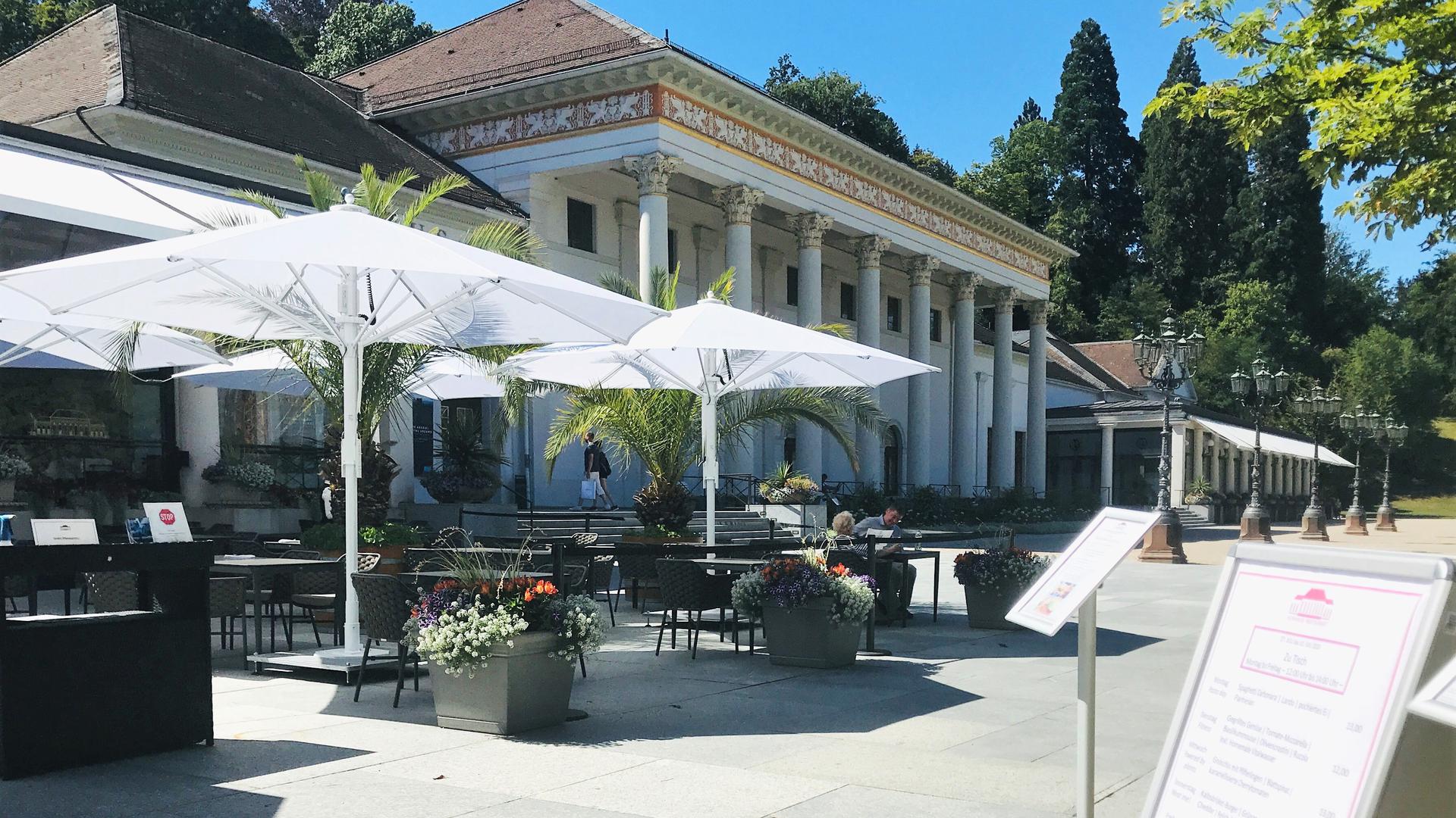 Vor einem Gebäude stehen Sonnenschirme und Tische und Stühle. Zu sehen ist die Terrasse des Kurhausrestaurants in Baden-Baden