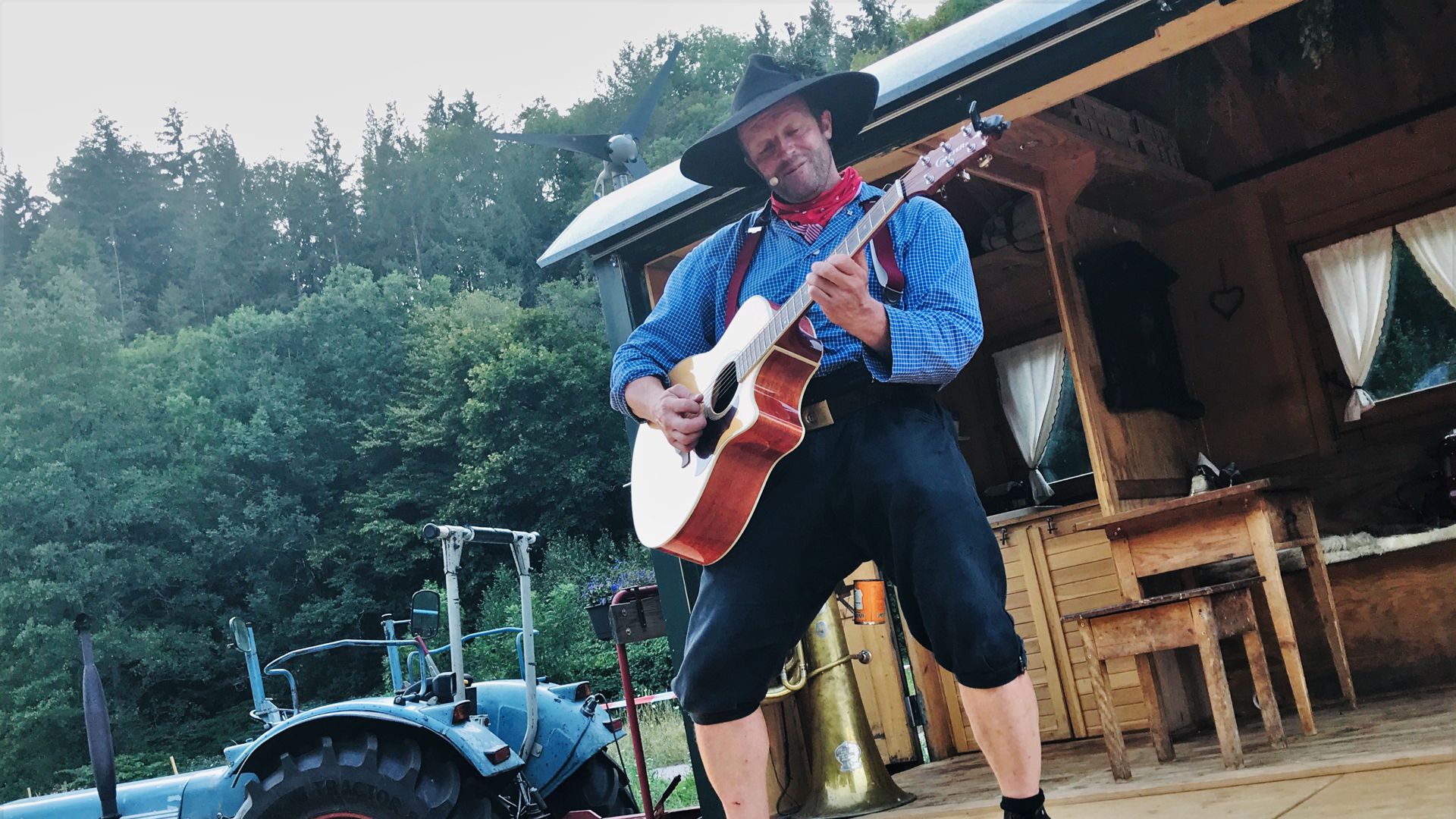 Ein Mann steht mit einer Gitarre auf einer Holzbühne. Es ist eine ausgeklappte Seitenwand eines umgebauten ehemaligen Forstwagens.