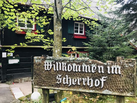 Vor einem Holzgebäude weist ein Schild mit der Aufschrifft „Willkommen im Scherrhof“ auf das Ausflugslokal im Stadtwald Baden-Baden hin. 