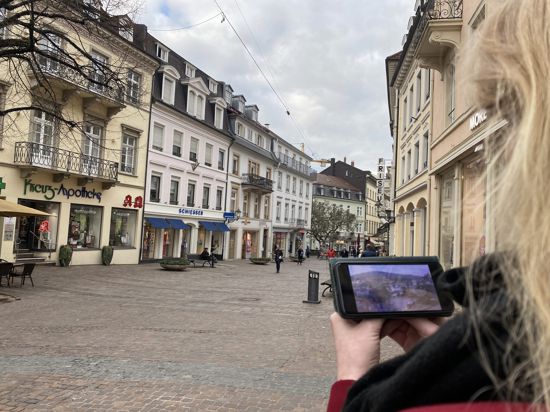 Eine Frau steht in der Innenstadt von Baden-Baden und schaut auf ihr Smartphone, das ein Video einer Webcam vom Goetheplatz anzeigt.