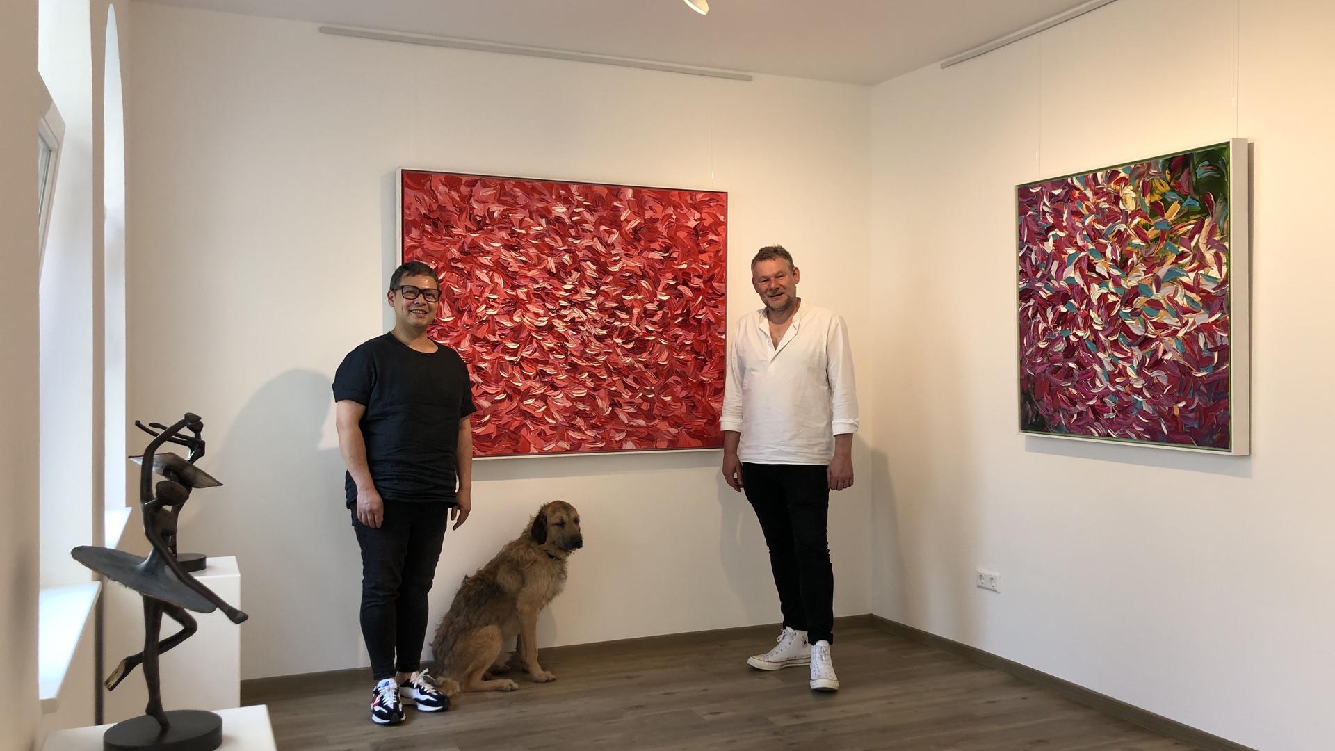 Zwei Männer und ein Hund in einer Galerie vor zwei Gemälden.