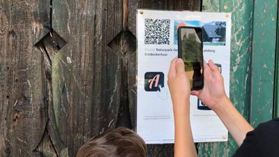 Zwei Kinder scannen mit einem Smartphone einen QR-Quode an einer Holzwand