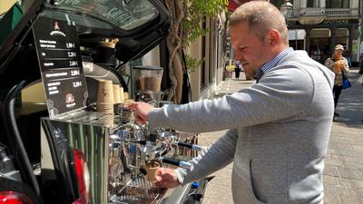 Ein Mann bereitet an einer mobilen Espresso-Bar im Kofferraum eines Kleinwagens einen Kaffee zu.