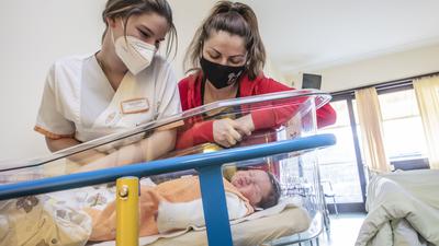 Zwei Krankenschwestern beugen sich über ein Bettchen, in dem ein Baby liegt.