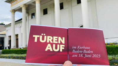 Vor dem Kurhaus hält eine Hand einen Flyer zum Tag der offenen Tür am 18. Juni im Kurhaus in Baden-Baden.
