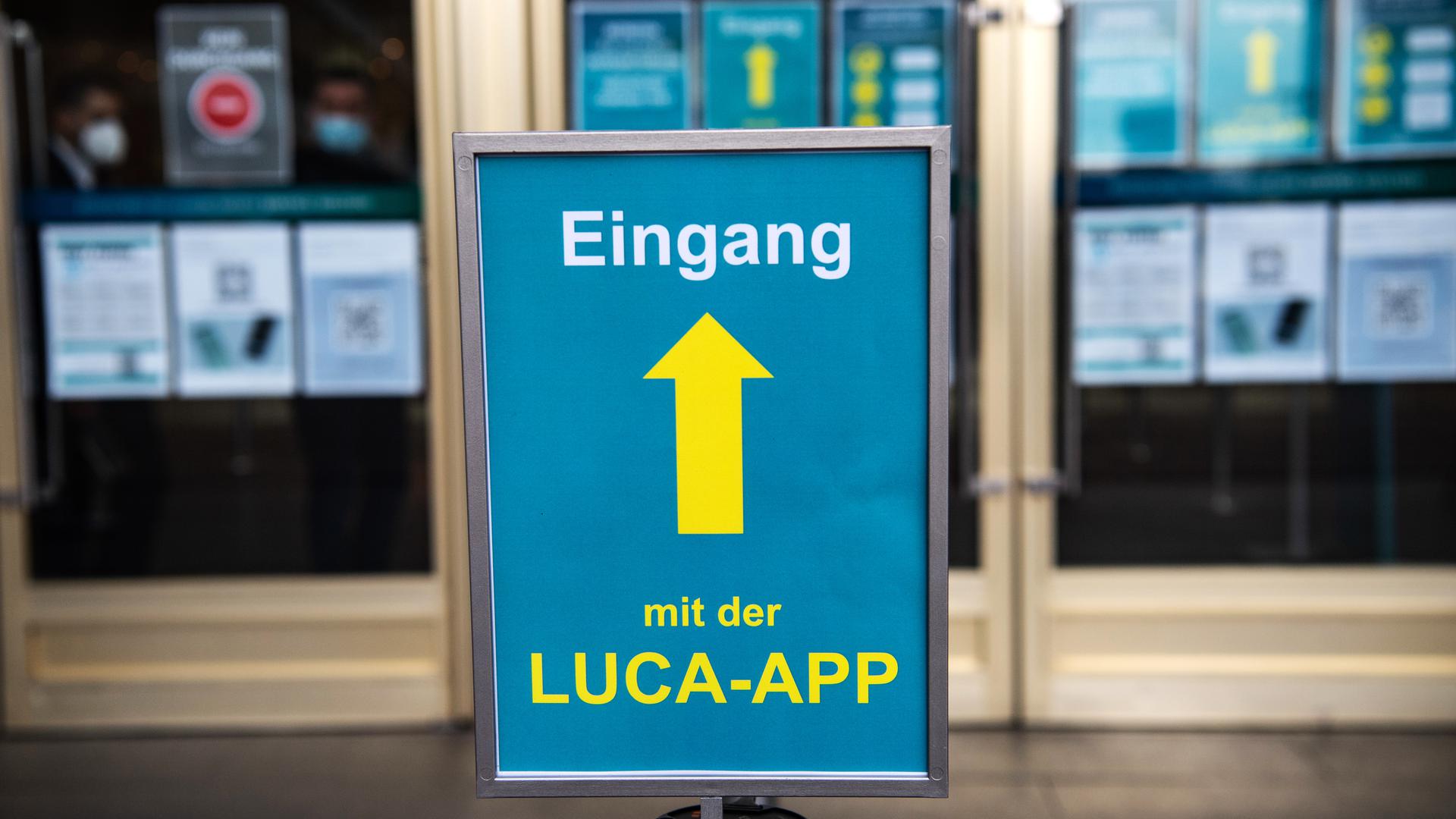 Ein blaues Schild mit einer gelben Aufschrift „Eingang mit der Luca-App“ steht vor einem Türeingang eines Gebäudes.