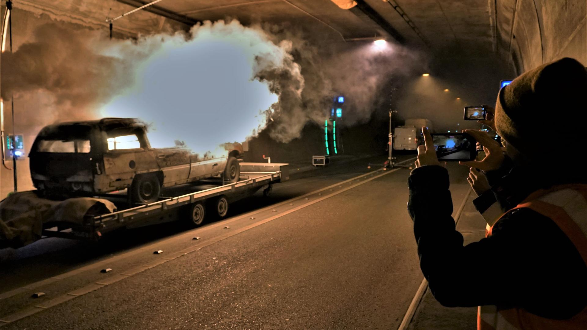 In einem Tunnel brennt ein Autowrack. Menschen halten das Ereignis mit ihren Handys fest.