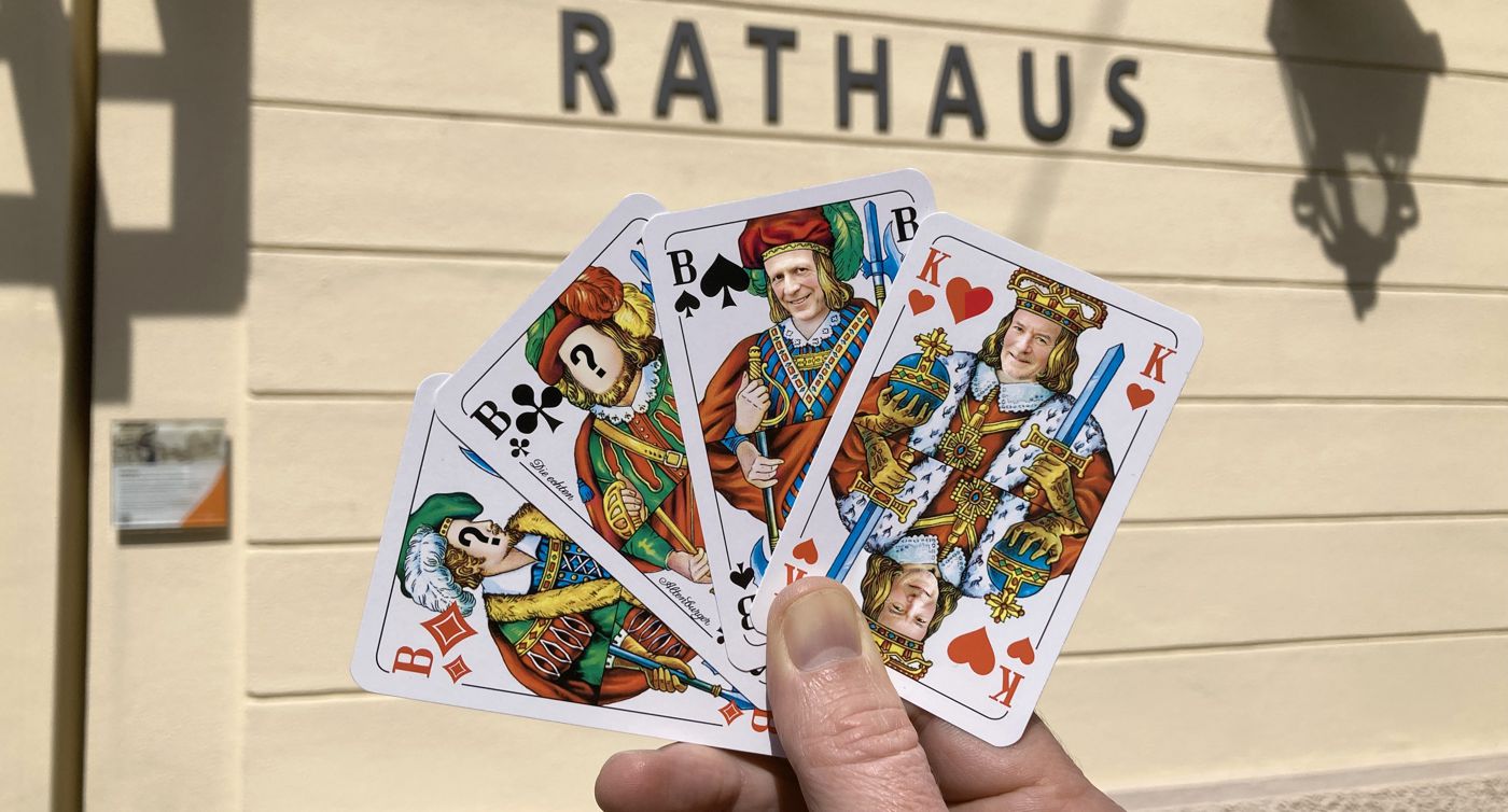 Eine Hand hält Skatkarten mit drei Buben und einem König vor dem Baden-Badener Rathaus. Auf den König ist das Gesicht von OB Dietmar Späth montiert, auf einen Bube das von Bürgermeister Roland Kaiser.