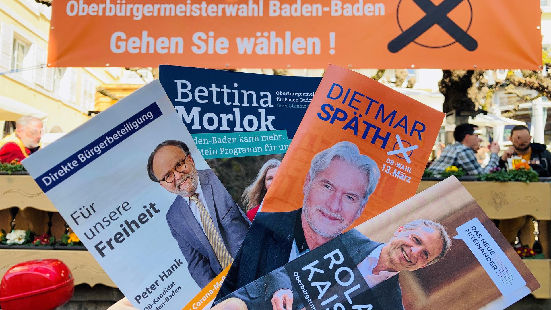Vor einem Transparent zur Oberbürgermeisterwahl in Baden-Baden werden Flyer der vier Bewerber gehalten. 
