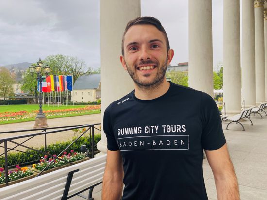 Vor dem Kurhaus in Baden-Baden steht ein junger Mann in einem schwarzen T-Shirt mit der Aufschrift „Running City Tours“. 