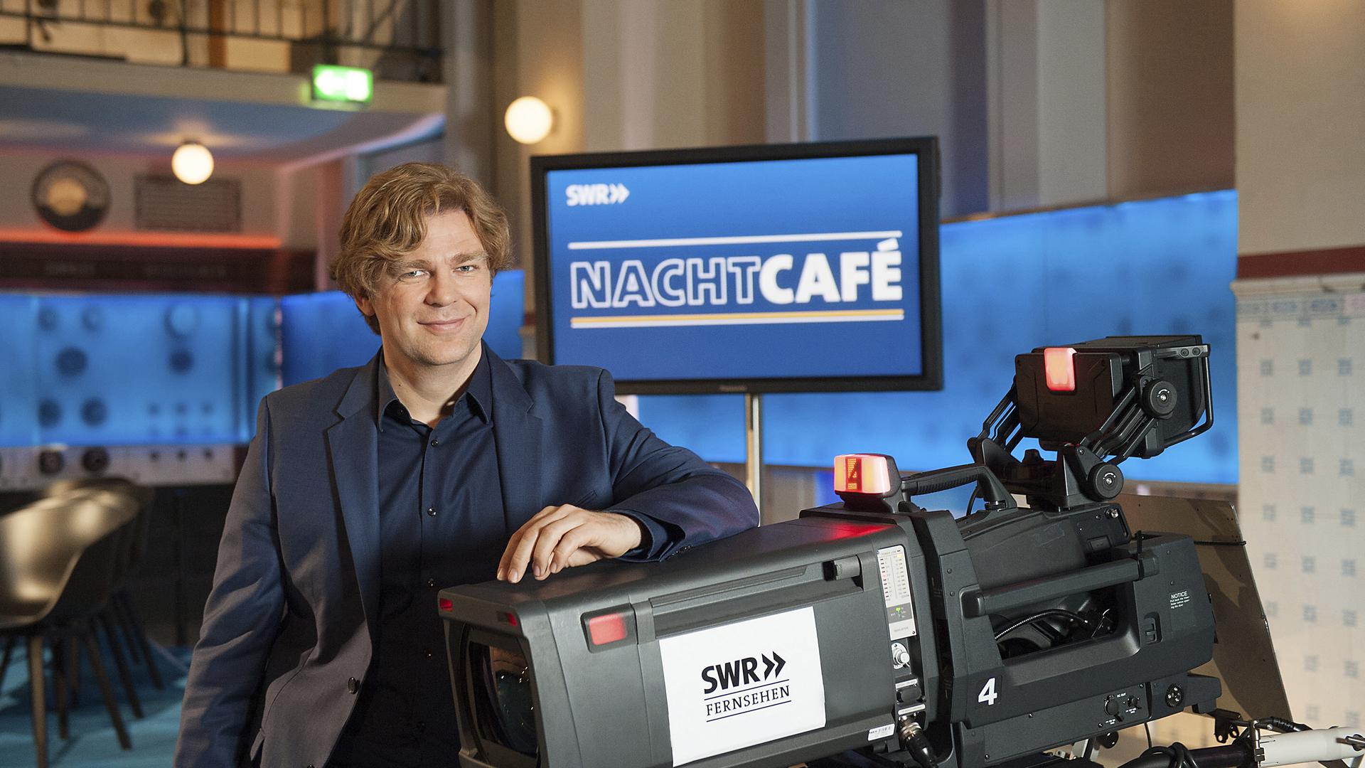 Michael Steinbrecher, Moderator der SWR-Talkrunde „Nachtcafé“ steht an einer Fernsehkamera.