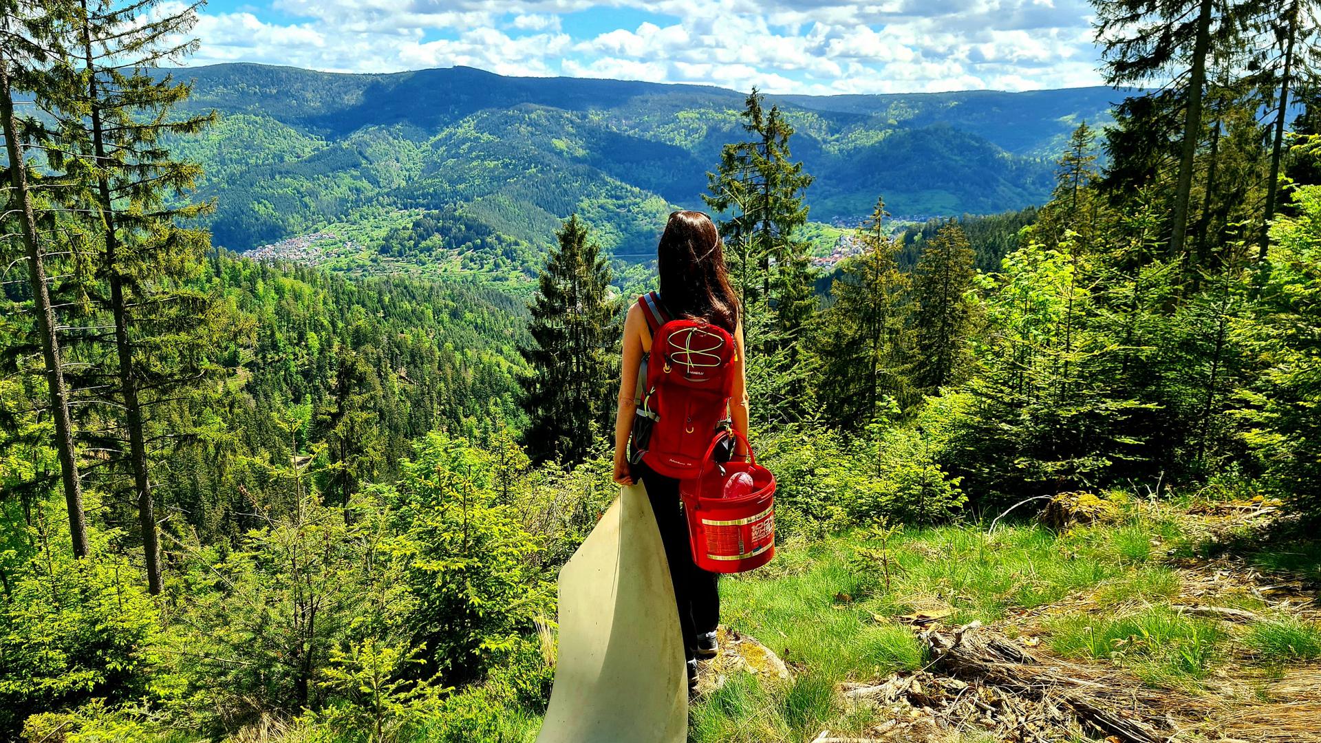 Frau steht vor Aussicht in der Natur und hält Plastikplane fest. Ein roter Eimer baumelt an ihrem Rucksack.