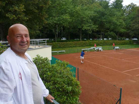 Ein Mann lacht in die Kamera, im Hintergrund sind Tennisspieler zu sehen