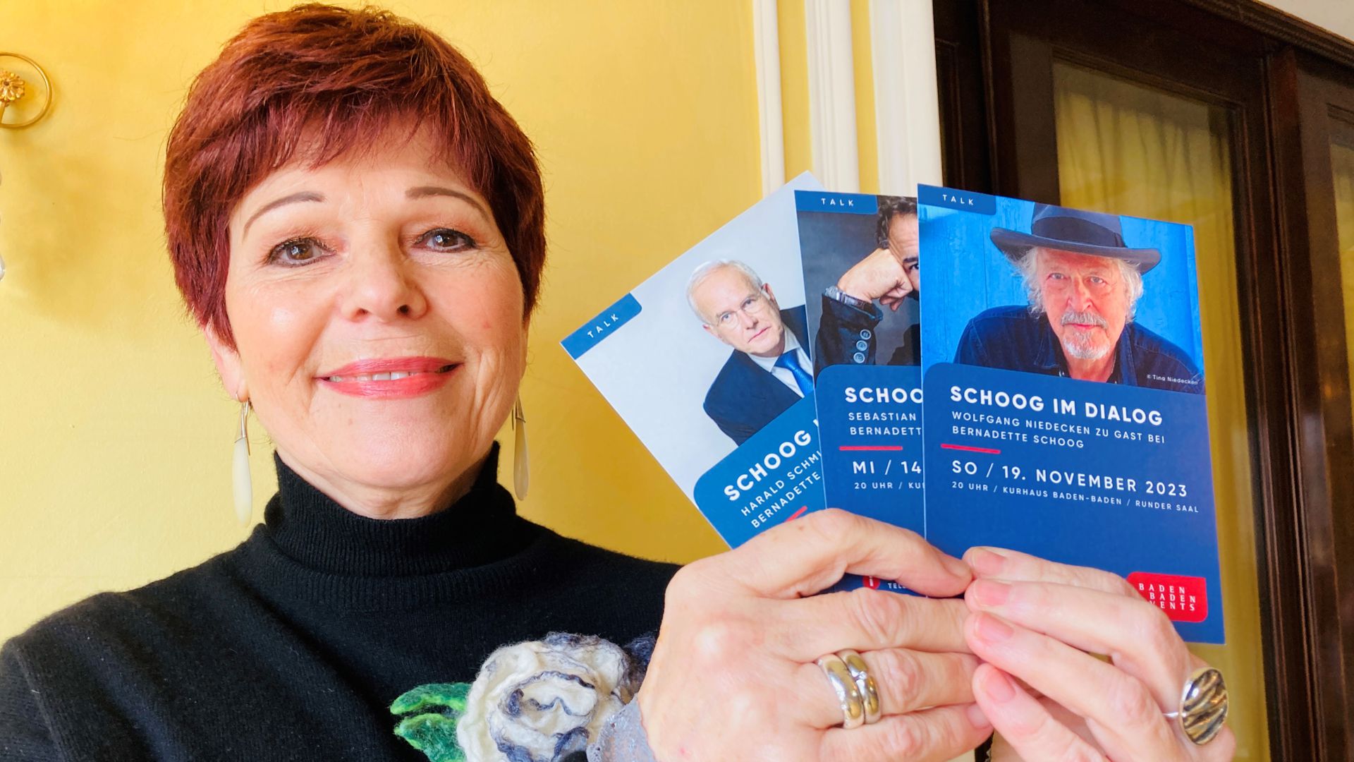 Moderatorin Bernadette Schoog hält Werbe-Karten für ihre Reihe „Schoog im Dialog“ in der Hand. 