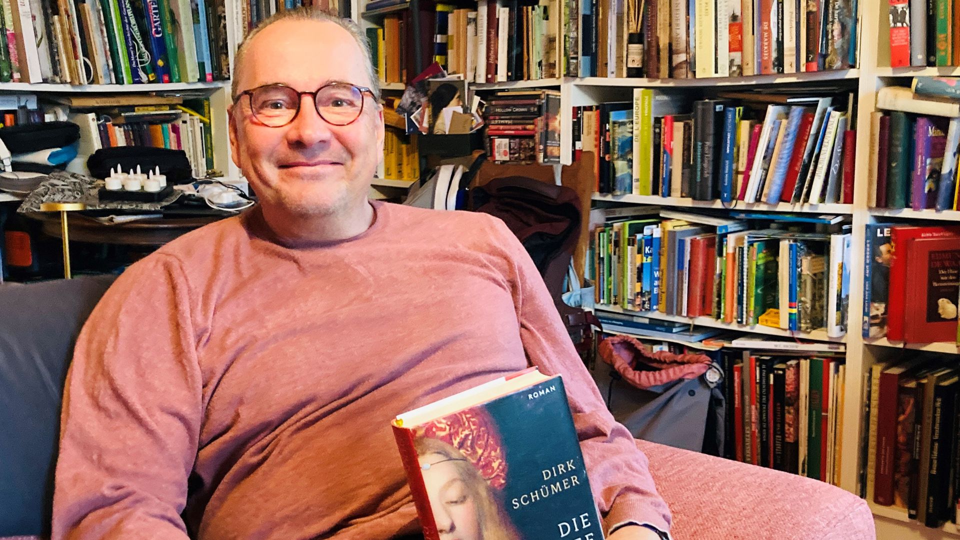 Autor Dirk Schümer sitzt mit seinem Roman „Die schwarze Lilie“ auf einem Sofa.