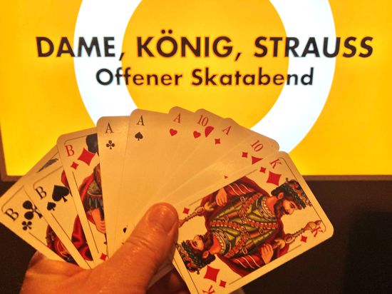 Eine Hand hält Skatkarten vor einem Leuchtdisplay mit der Aufschrift „Dame, König, Strauss“. 