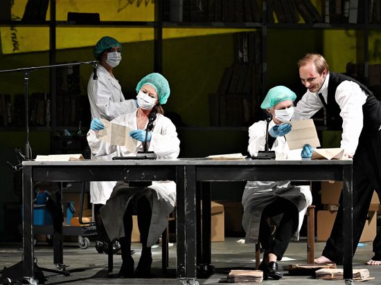 Mehrere Schauspielerinnen mit Mund-Nasen-Schutz sitzen in dem Stück „Der Fall Hau“ des Theaters Baden-Baden an einem Tisch auf der Bühne.