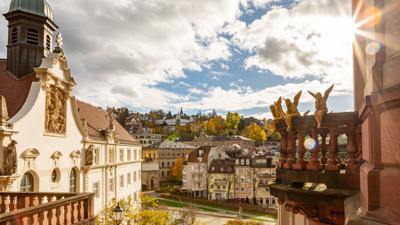 Blick über Baden-Baden vom Bäderviertel aus, am Bildrand sieht man einen Balkon mit goldenen Engeln. Dahinter bricht die Sonne durch die Wolken.