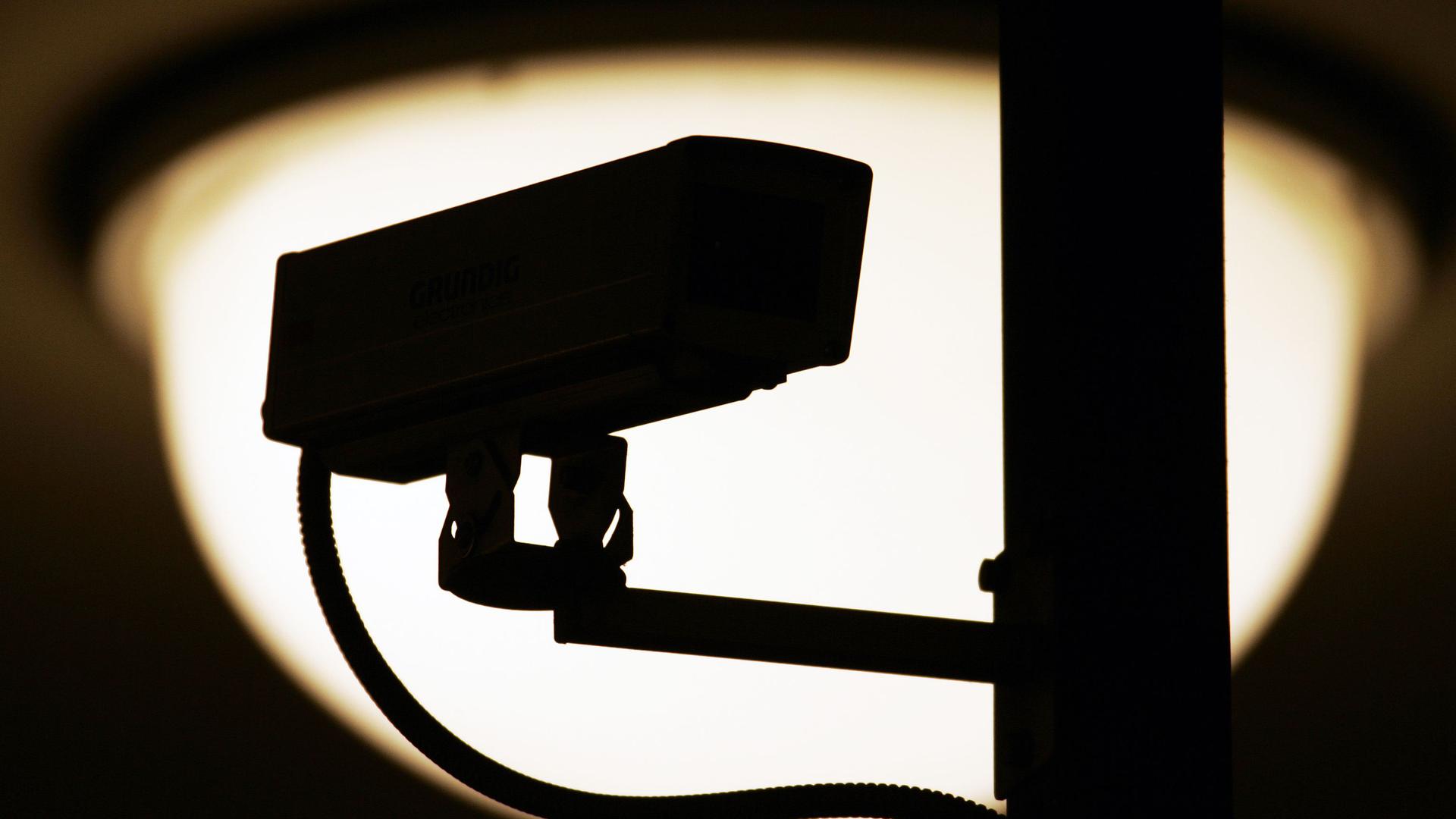 Eine Video-Überwachungskamera zeichnet sich vor einer Lampe als dunkle Silhouette ab.