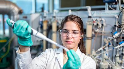 Eine Frau in Laborkleidung pipettiert etwas. Im Hintergrund eine Industrieanlage.