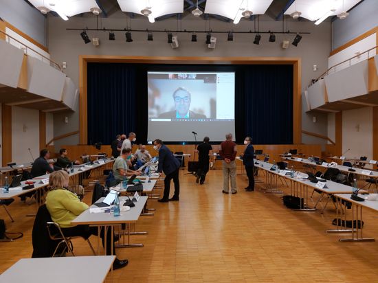 Innenraum einer Halle, in der eine Gemeinderatssitzung stattfindet. Auf einer Leinwand ist das Gesicht eines Mannes zu sehen, der digital zugeschaltet ist.