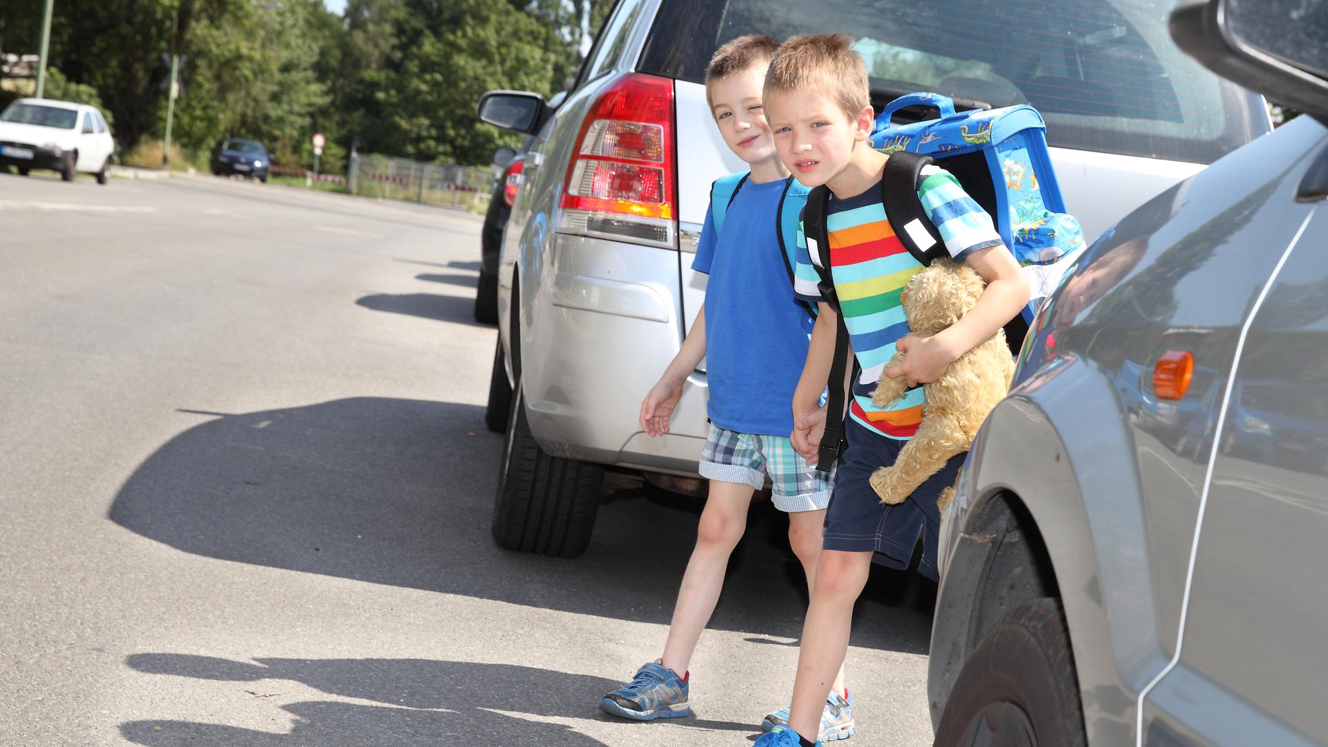 Zwei Jungen wollen hinter einem parkenden Auto stehend über die Straße gehen.