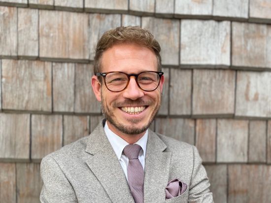 Ein Mann mit Brille, kurzem Vollbart und hellem Anzug mit violetter Krawatte und Einstecktuch lächelt breit in die Kamera