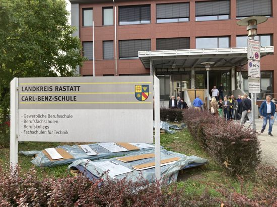 im Vordergrund: ein Schild mit der Aufschrift „Landkreis Rastatt - Carl-Benz-Schule“, dahinter Baumaterial auf einer Wiese. Im Hintergrund: die Fassade eines Gebäudes und der unfertige Haupteingang