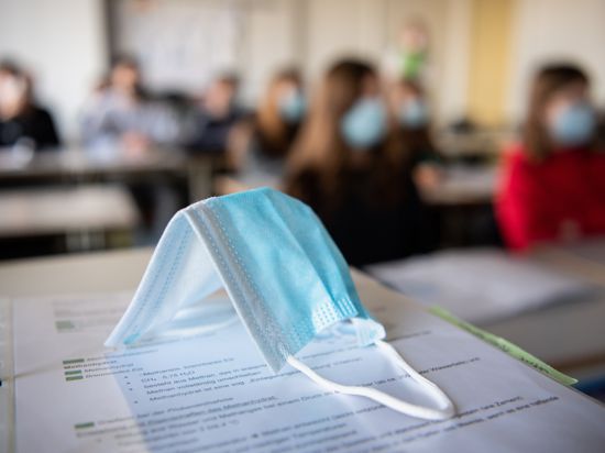 ILLUSTRATION - Eine Maske liegt im Unterricht in einem Gymnasium auf Unterrichtsunterlagen, während im Hintergrund Schülerinnen und Schüler mit Mund- und Nasenschutz zu sehen sind. (Zu dpa «Länder-Entwurf: Maskenpflicht in Corona-Hotspots ab Klasse sieben»)