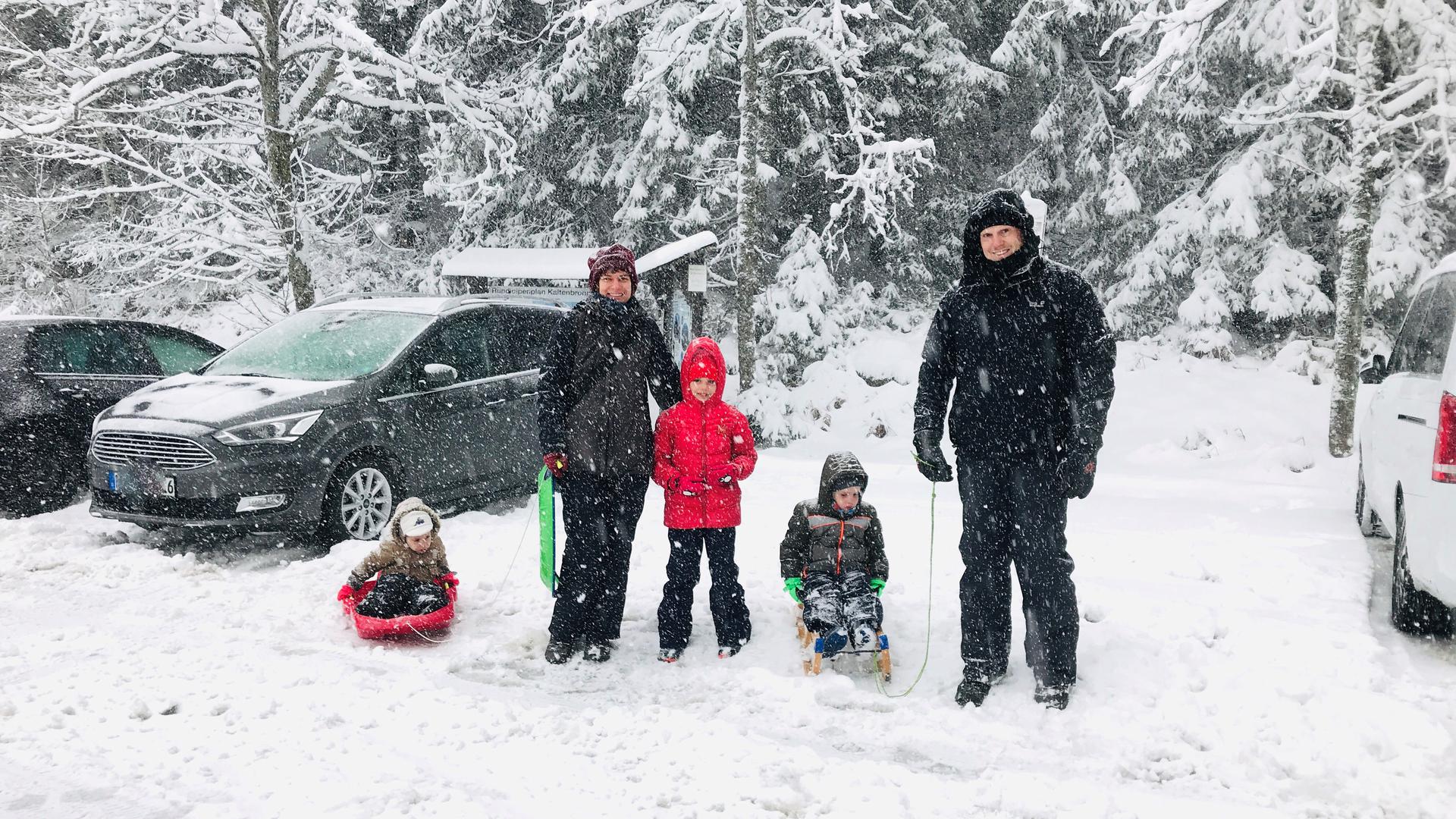 Eine Familie mit drei Kindern steht mit Rodeln auf dem schneebedeckten Parkplatz bei Kaltenbronn