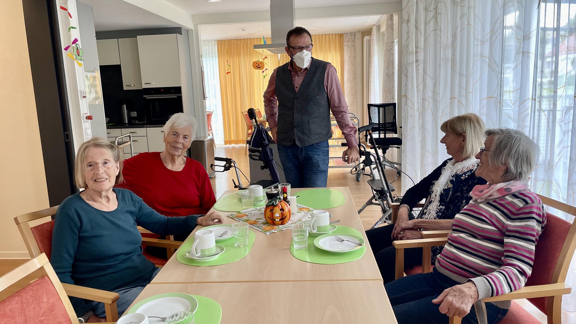 Alte Leute sitzen um einen Tisch, im Hintergrund ein stehender Mann mit Maske