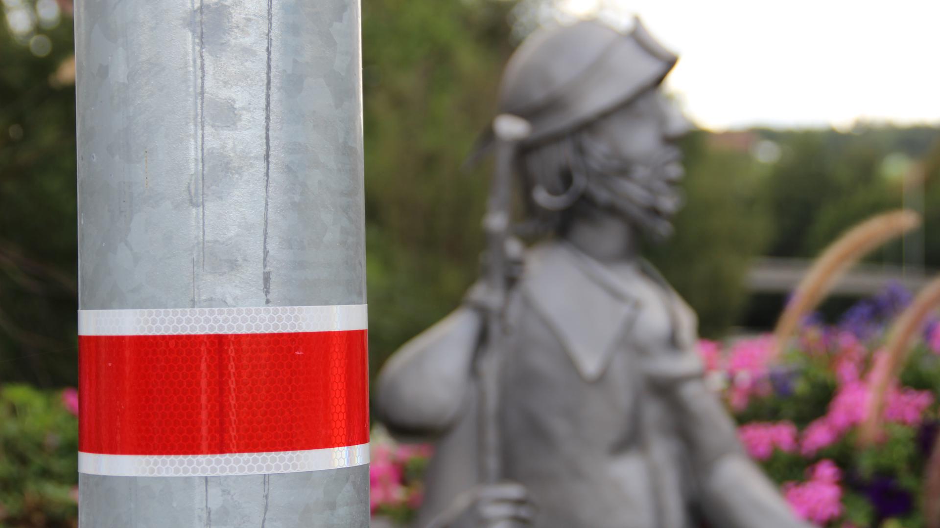 Mast einer Straßenlaterne mit dem Verkehrszeichen 394 „Laternenring“. Es ist der Hinweis, dass die Laterne nicht die ganze Nacht hindurch brennt. Im Hintergrund die Figur des Jakobspilgers auf der Konrad-Adenauer-Brücke in Gaggenau.