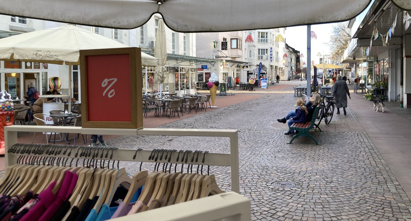 Ein Kleiderständer in der Gaggenauer Fußgängerzone. Dahinter der Blick auf die Hauptstraße mit Menschen, die auf Bänken sitzen. 