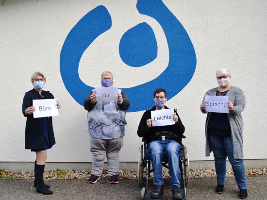 Vor einer Wand mit dem blauen Logo der Lebenshilfe zeigen vier Personen Schilder mit dem Hinweis „Büro für leichte Sprache“