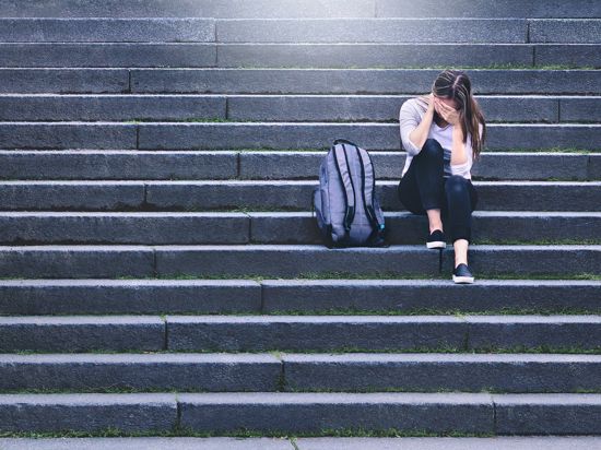 Bullying, discrimination or stress concept. Sad teenager crying in school yard. Upset young female student having anxiety. Upset victim of abuse or harassment sitting on stairs outdoors with backbag.