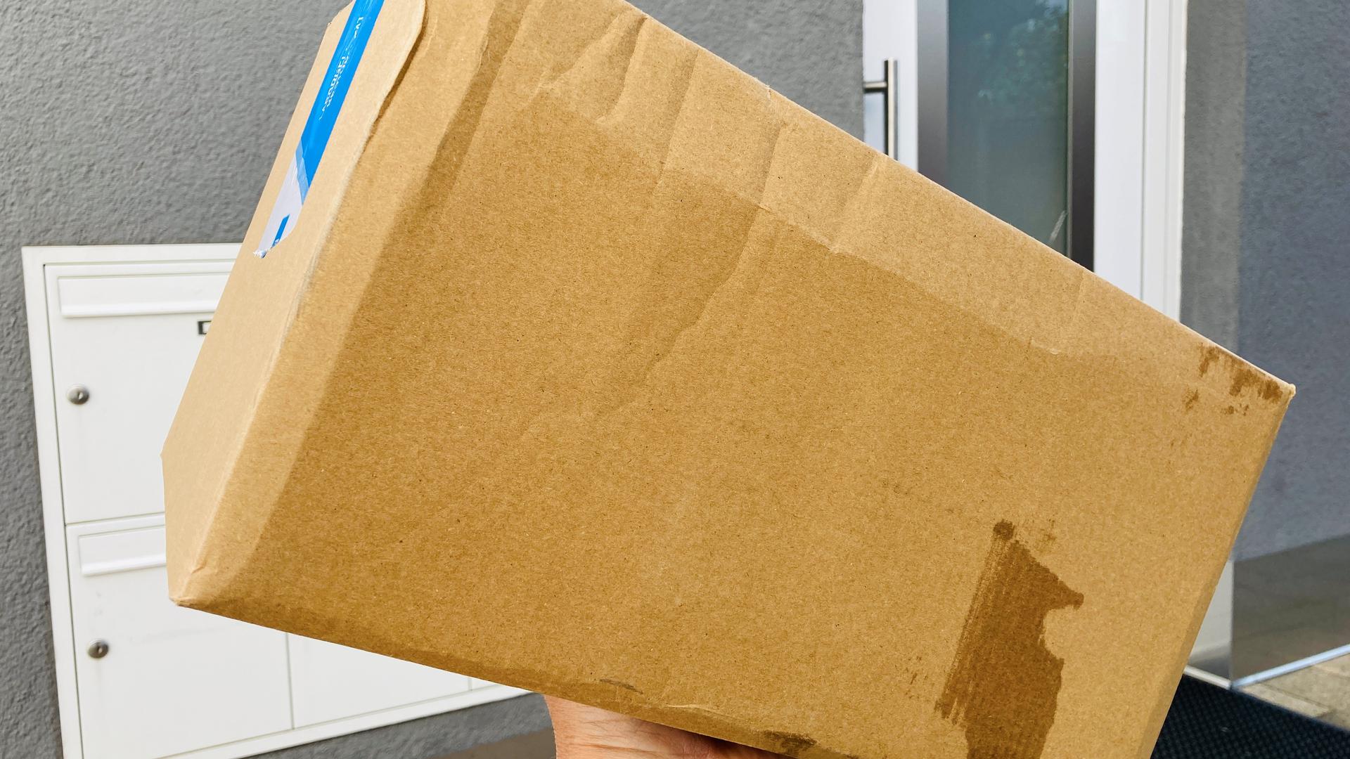 Eine Hand hält ein Paket vor einer Haustür mit einer Briefkastenanlage.