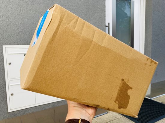 Eine Hand hält ein Paket vor einer Haustür mit einer Briefkastenanlage.