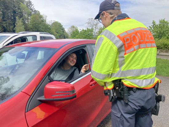 ein Polizist beugt sich bei einer Verkehrskontrolle zum geöffneten Fenster eines roten Autos und reicht der Fahrerin ihre Papiere 
