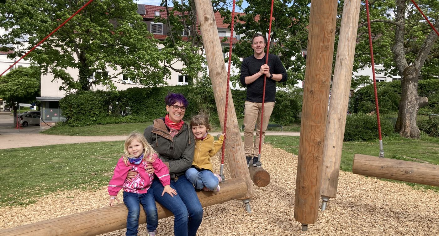 zwei Kinder und zwei Erwachsene sitzen und stehen auf einer großen Holzkonstruktion im Freien, bei der Balken über Seile miteinander verbunden sind und schwingen können